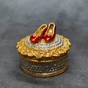 May include: A small, round jewelry box with a gold and silver finish. Two red, high-heeled shoes with gold accents sit atop the lid. The box has a detailed, ornate design, perfect for storing small items.