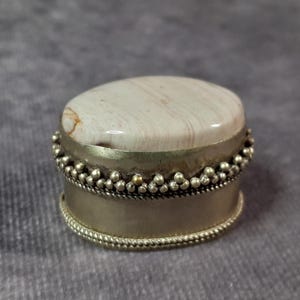 May include: A small, round trinket box with a light-colored, polished stone top. The box has a silver-colored metal band with a beaded design around the middle and bottom. The stone top has brown and tan veining.
