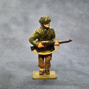 Op de afbeelding: Een beschilderd metalen speelgoed soldaat figuur. De soldaat draagt een groene camouflagehelm en jas, bruine broek en houdt een bruin geweer met een gele riem vast. Het beeldje staat op een kleine, rechthoekige basis.