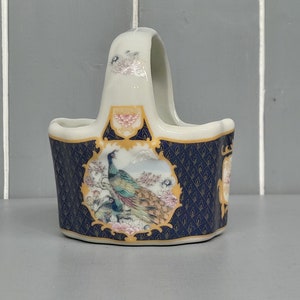 May include: A small white ceramic basket with a handle, decorated with a blue and gold floral pattern and a peacock design.