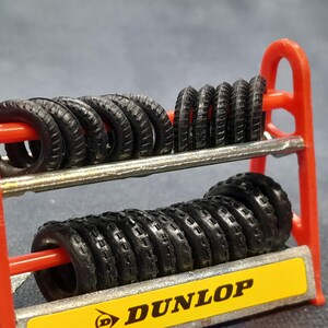 K&ouml;nnte beinhalten: Ein rotes Reifenregal mit mehreren schwarzen Reifen. Das Regal hat zwei Ebenen und ein gelbes Schild mit dem Wort "DUNLOP". Die kleinen Reifen haben ein detailliertes Profil.