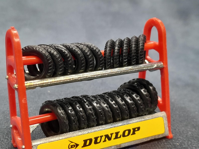 K&ouml;nnte beinhalten: Ein rotes Reifenregal mit mehreren schwarzen Reifen. Das Regal hat zwei Ebenen und ein gelbes Schild mit dem Wort "DUNLOP". Die kleinen Reifen haben ein detailliertes Profil.