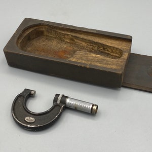 Könnte beinhalten: Ein schwarzer und silberner Mikrometer mit einem Holzkasten. Der Mikrometer hat eine Skala mit Zahlen von 0 bis 10. Der Kasten ist aus dunklem Holz und hat einen Klappdeckel.
