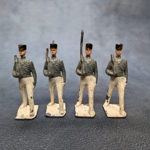 Puede incluir: Cuatro soldados de juguete antiguos en uniforme. Cada figura lleva una chaqueta gris, pantalones blancos y un sombrero negro. Los soldados sostienen rifles u otro equipo militar. Las figuras están sobre bases rectangulares.