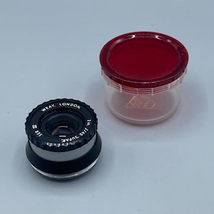 Pu&ograve; includere: Un obiettivo per macchina fotografica nero e argento con la scritta "WRAY. LONDON 2 in. f/1.5 SUPAR" stampata sul lato. L'obiettivo &egrave; appoggiato su una superficie bianca accanto a un contenitore di plastica rosso e trasparente.