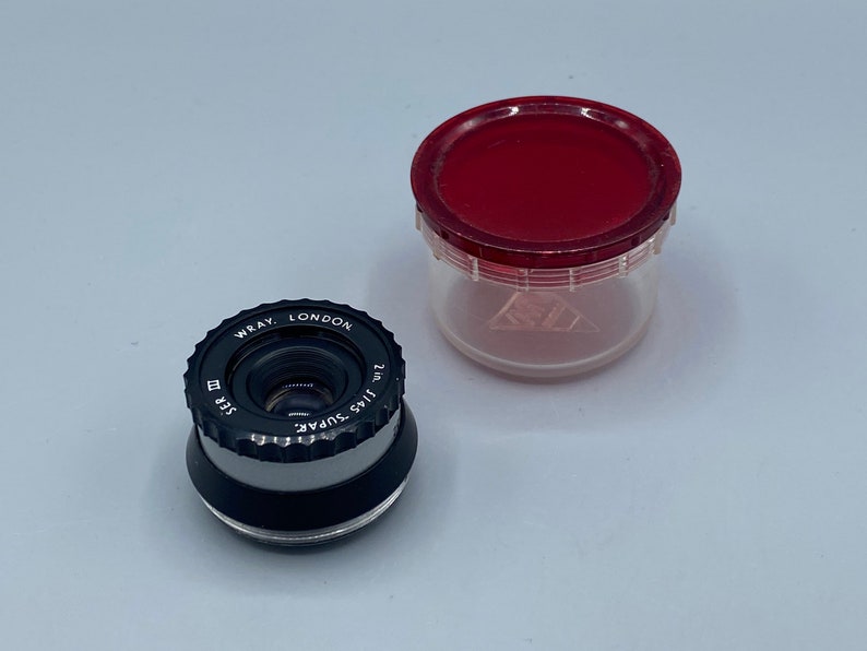 Pu&ograve; includere: Un obiettivo per macchina fotografica nero e argento con la scritta "WRAY. LONDON 2 in. f/1.5 SUPAR" stampata sul lato. L'obiettivo &egrave; appoggiato su una superficie bianca accanto a un contenitore di plastica rosso e trasparente.