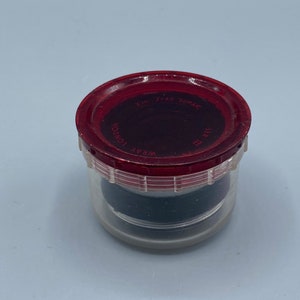 Pu&ograve; includere: Un contenitore di plastica trasparente con un coperchio rosso. Il coperchio ha la scritta "WRAY LONDON" e "FIRST SURFACE" stampata. Il contenitore &egrave; riempito con un oggetto nero.