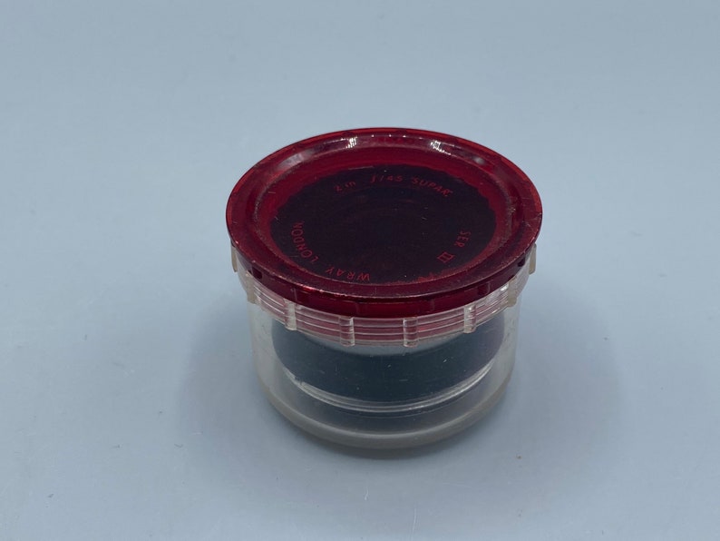 Pu&ograve; includere: Un contenitore di plastica trasparente con un coperchio rosso. Il coperchio ha la scritta "WRAY LONDON" e "FIRST SURFACE" stampata. Il contenitore &egrave; riempito con un oggetto nero.