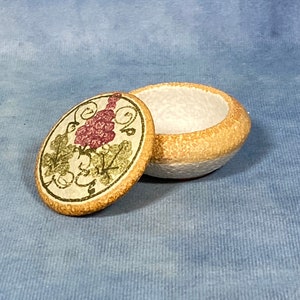 Peut inclure: Une petite boîte à bijoux ronde en céramique avec un couvercle. Le couvercle est décoré d'un motif peint de vigne avec des feuilles vertes et des raisins violets. La boîte est de couleur brun clair avec un intérieur blanc.