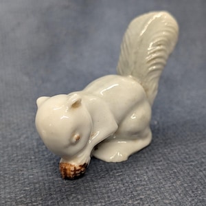 May include: A small, glossy white ceramic figurine of a squirrel in a seated position. The squirrel has a large, curved, textured tail and is holding a small, brown, textured nut between its front paws, with its head bowed downward.
