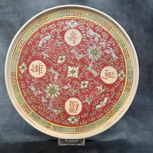 Peut inclure: Une assiette décorative ronde avec un centre rouge orné de motifs floraux et de volutes blancs. L'assiette comporte quatre caractères chinois dans des cercles et un bord crème avec un motif géométrique doré et vert. L'assiette est sur un support transparent.