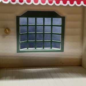 May include: A miniature dollhouse interior with a white interior and a green-framed window. The window has a grid pattern. The roof is red with a scalloped edge. A small, round, yellow object is on the wall.