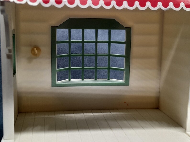 May include: A miniature dollhouse interior with a white interior and a green-framed window. The window has a grid pattern. The roof is red with a scalloped edge. A small, round, yellow object is on the wall.