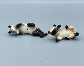May include: Two small ceramic Siamese cat figurines, both lying on their backs with their paws in the air. One cat is white with black markings, and the other is beige with black markings.