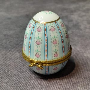 Puede incluir: Una caja de baratijas en forma de huevo con tapa con bisagras. La caja es de color azul claro con un patrón repetido de formas florales rosas y hojas verdes. Los adornos dorados acentúan los bordes y la bisagra.