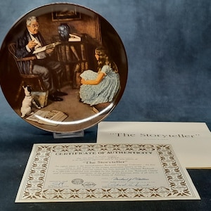 May include: A decorative plate with the image "The Storyteller", depicting a man reading to a girl and a dog. Includes a certificate of authenticity and a paper with the title. The plate is a decorative item.