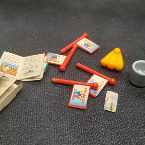 May include: A collection of miniature Sylvanian Families accessories, including red flag signs with the text "Sylvanian Games", a small yellow cone, a gray cup, and a booklet with illustrations.