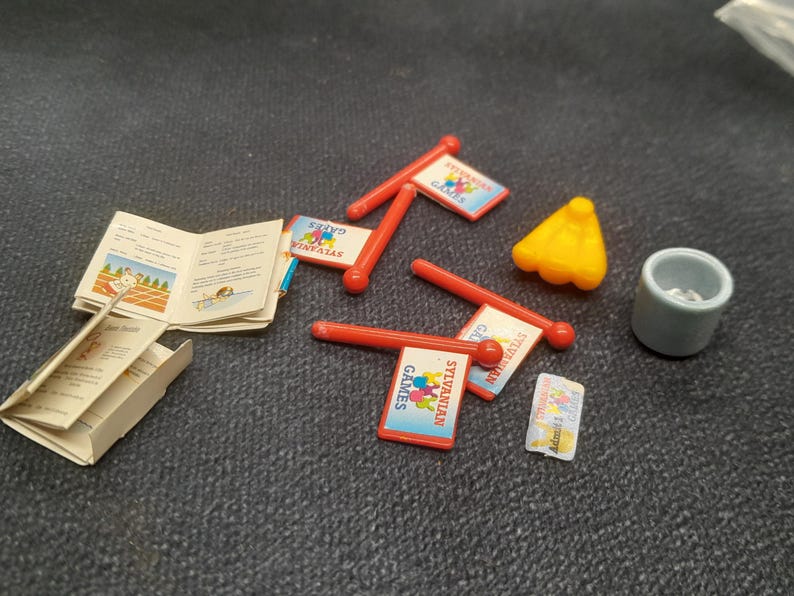 May include: A collection of miniature Sylvanian Families accessories, including red flag signs with the text "Sylvanian Games", a small yellow cone, a gray cup, and a booklet with illustrations.