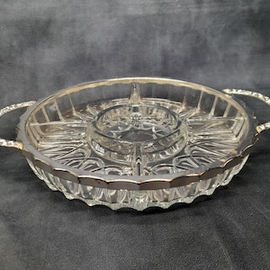 May include: A clear glass serving tray with a silver-coloured rim and handles. The tray is divided into sections and has a central circular compartment. The handles have a textured design.