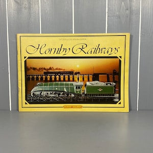 以下が含まれることがあります： 「Hornby Railways」というタイトルが大きく太字で印刷された黄色と茶色の本。表紙には、緑と黒の模型列車が橋を渡っている写真が掲載されています。