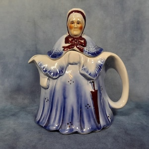 以下が含まれることがあります： 青いドレスと帽子を着た女性の形をしたセラミックティーポット。 ティーポットは白いハンドルと注ぎ口があり、マルーンのリボンとアクセントが付いています。 デザインには、青い花のディテールとマルーンの傘が含まれています。