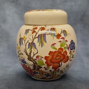 May include: Cream-coloured ceramic jar with a lid, decorated with a floral and tree design. The jar features a large orange flower, blue and purple flowers, and a tree with red berries. The lid has a matching floral pattern.