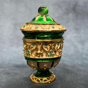 Puede incluir: Un tarro decorativo y ornamentado con tapa. El tarro es principalmente verde y dorado, con una banda color crema en el medio. La tapa está rematada con una piedra verde. El tarro tiene una base de pedestal y probablemente es de metal.