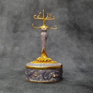 Könnte beinhalten: Ein dekorativer Schmuckhalter mit goldfarbenem Oberteil und Sockel sowie einem grau-goldfarbenen Körper. Der Schmuckhalter hat einen runden Sockel mit einem Wirbeldesign und ist mit klaren Edelsteinen verziert. Die Oberseite hat mehrere Haken zum Aufhängen von Schmuck.