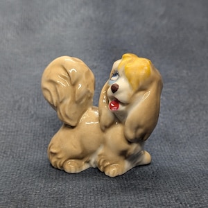 May include: A small ceramic figurine of a tan dog with long, flowing ears and a bushy, upright tail. The dog has a yellow patch of fur on its head, blue painted eyes, a dark brown nose, and a small red tongue visible in its open mouth.