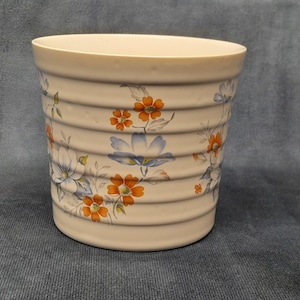 May include: A white ceramic planter with a floral design featuring blue and orange flowers. The planter has a ribbed texture and a slightly rounded shape.
