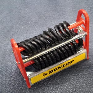 K&ouml;nnte beinhalten: Ein Miniatur-Reifenst&auml;nder mit schwarzen Reifen, einem roten Rahmen und einer gelb-silbernen Basis. Die Basis zeigt das Dunlop-Logo. Die Reifen sind in Reihen gestapelt, was auf ein Modellauto-Zubeh&ouml;r hindeutet.