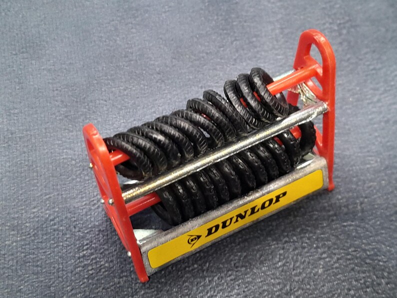K&ouml;nnte beinhalten: Ein Miniatur-Reifenst&auml;nder mit schwarzen Reifen, einem roten Rahmen und einer gelb-silbernen Basis. Die Basis zeigt das Dunlop-Logo. Die Reifen sind in Reihen gestapelt, was auf ein Modellauto-Zubeh&ouml;r hindeutet.
