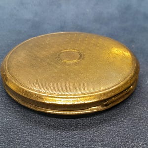 May include: A vintage, round, gold-toned compact with a textured surface. The lid features a central, slightly raised circle and a detailed, patterned design. The compact is closed, showing its complete form and intricate detailing.