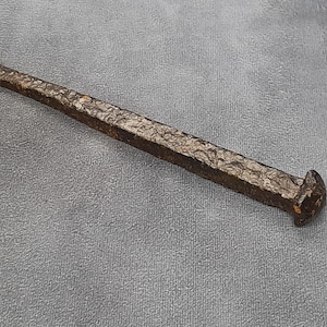 May include: An antique iron nail with a square head and a textured, rectangular shaft. The nail has a dark brown colour and appears to be hand-forged. The nail is lying on a grey surface.