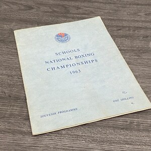 May include: A blue and white program for the Schools National Boxing Championships in 1963. The program is titled "Schools National Boxing Championships 1963" and says "Souvenir Programme" and "One Shilling" on the front.