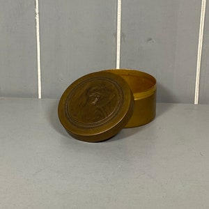 Könnte beinhalten: Eine runde Schatulle aus Messing mit einem erhabenen Design auf dem Deckel. Der Deckel ist geöffnet und die Schatulle ist leer.