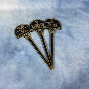 Op de afbeelding: Drie zwarte metalen tuinmarkeringen met gouden letters. De markeringen zijn gelabeld met "Bay", "Lavender" en "Chives".