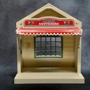 May include: A tan and white toy shop with a red awning and the words "GAMES SOUVENIRS" on a sign. The shop has a green-framed window with a grid pattern and a small gold-colored door handle.