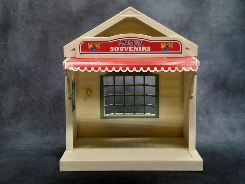 May include: A tan and white toy shop with a red awning and the words "GAMES SOUVENIRS" on a sign. The shop has a green-framed window with a grid pattern and a small gold-colored door handle.