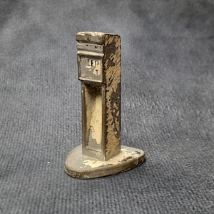 Può includere: Una piccola pompa di benzina in metallo in stile vintage. La pompa è dipinta nei toni del beige e del marrone, con segni di usura. Ha un corpo rettangolare e una base arrotondata. La parte superiore presenta un piccolo display rettangolare. Altezza circa 6 cm.