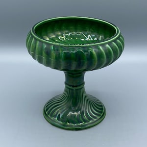 Może przedstawiać: Zielona ceramiczna miska na cokole z błyszczącym wykończeniem. Misa ma szeroką, żłobkowaną krawędź i spoczywa na szczegółowej, żebrowanej podstawie cokołu. Misa jest przeznaczona do celów dekoracyjnych.
