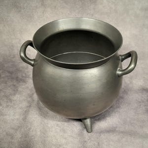 May include: A silver-colored cauldron with a rounded body, two side handles, and three small legs. The cauldron has a wide, flared rim and a dark interior. The metal pot is likely used for cooking or decorative purposes.