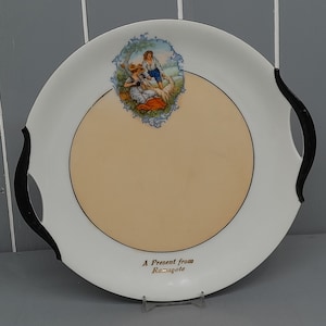 Puede incluir: Plato de servir de cerámica blanca con asas negras y borde dorado. En el centro del plato, una escena pintada que representa a dos personas en un campo con una oveja. El texto "A Present from Ramsgate" está impreso en dorado en la parte inferior del plato.