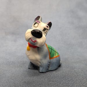 May include: A small ceramic dog figurine with a grey body, white muzzle, and black nose. The dog wears a red collar with a yellow tag and a green blanket with a black grid pattern on its back. The figurine features painted eyes and eyebrows.