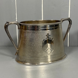 May include: A silver sugar bowl with a decorative embossed design and two handles. The bowl has a scalloped edge and a raised crest in the centre.
