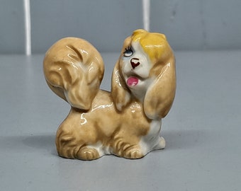 May include: A small ceramic figurine of a brown and white dog with yellow fur on its head. The dog has blue eyes and a pink tongue sticking out.