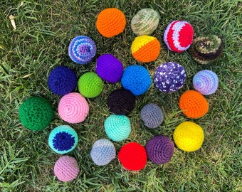 May include: A collection of colorful crocheted balls in various colors, including red, blue, green, yellow, and purple. The balls are arranged in a circle on a bed of green grass.