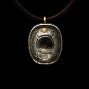 Puede incluir: Un colgante plateado con forma de balón de fútbol, suspendido de un cordón de cuero marrón. El colgante tiene una superficie detallada y texturizada y está sujeto al cordón con un pequeño lazo dorado. El fondo es negro.