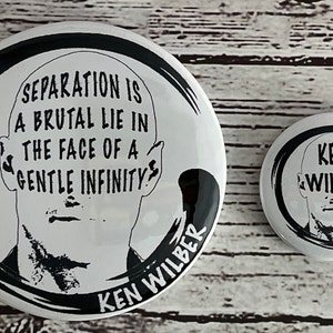 May include: Two white round buttons with black ink. The larger button has a black and white illustration of a person's head with the text "SEPARATION IS A BRUTAL LIE IN THE FACE OF A GENTLE INFINITY" and "KEN WILBER". The smaller button has a black and white illustration of a person's head with the text "KEN WILBER".