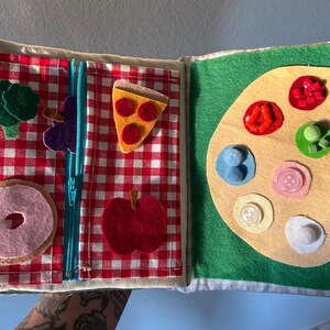 May include: A felt book with two pages. The left page has a red and white checkered pattern with felt images of a pizza slice, a donut, a bunch of broccoli, a grape, and an apple. The right page has a green background with a felt image of a paint palette with different colored felt shapes.